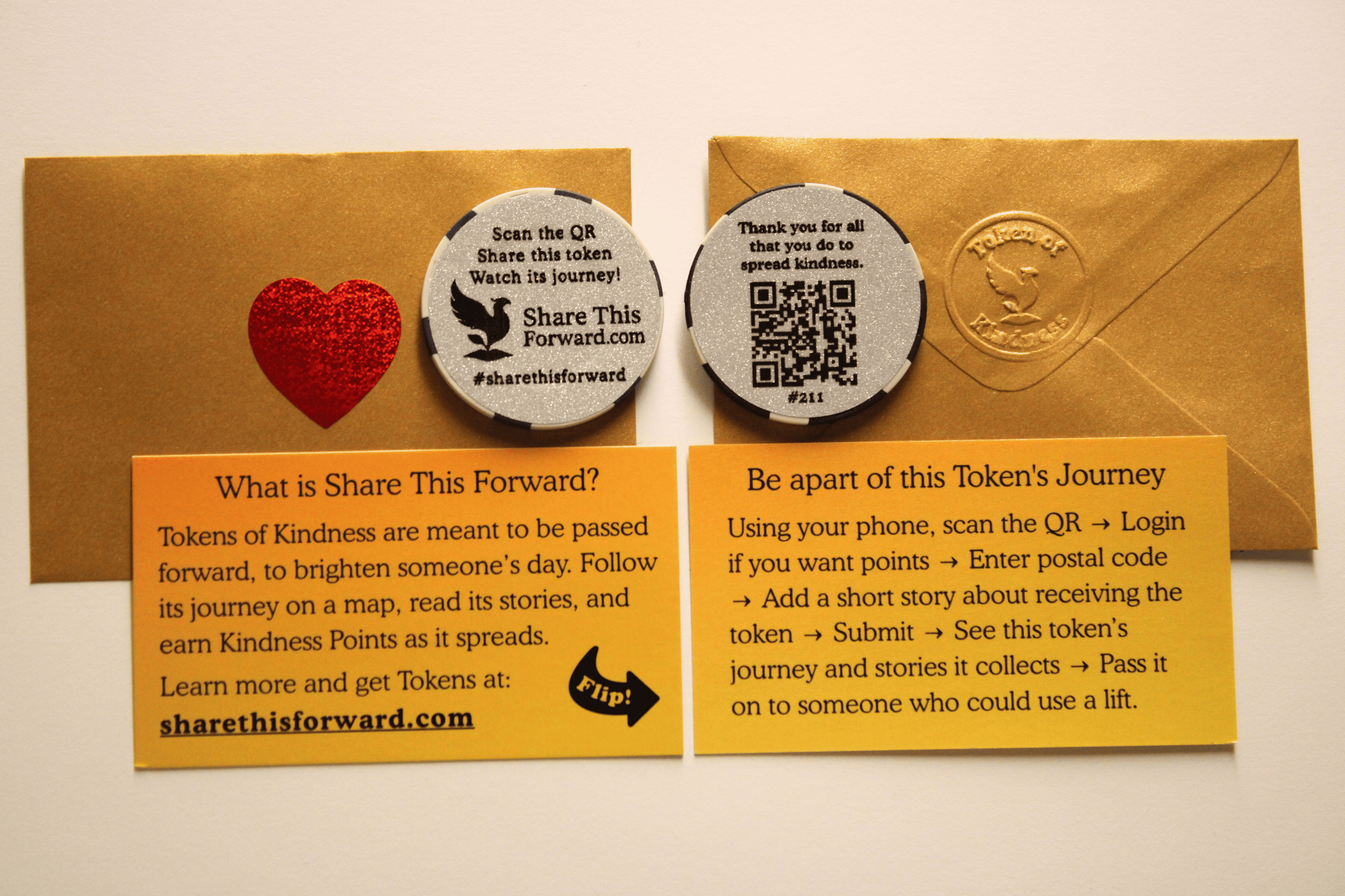 A Share This Forward Kindness Token displayed with its gold envelope, heart sticker, and instruction cards. The token features a QR code that lets people track its journey and share acts of kindness as it’s passed forward.